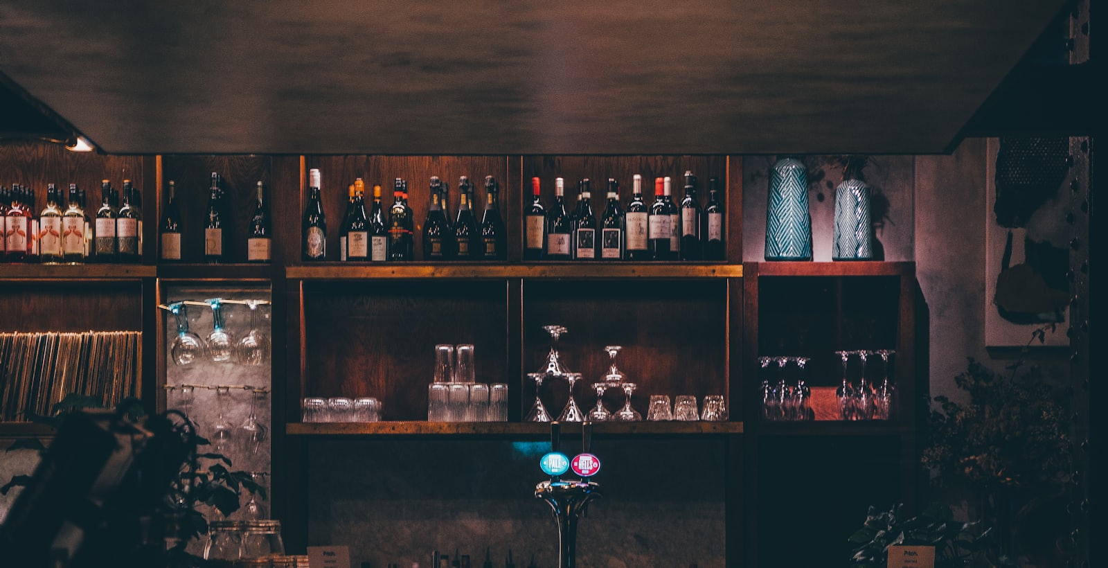 A low-lit bar back-counter — shelves of bottles and glassware glowing in warm light